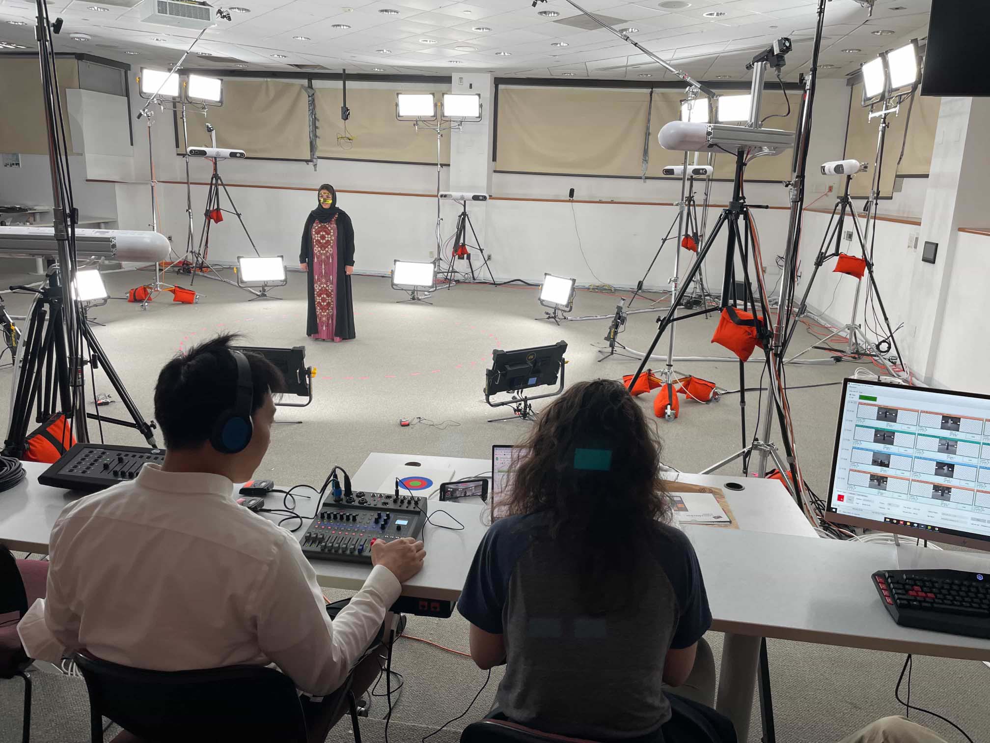 Two people sit and observe a woman dressed in traditional Emirati clothing stands at the center of a volumetric capture studio stage