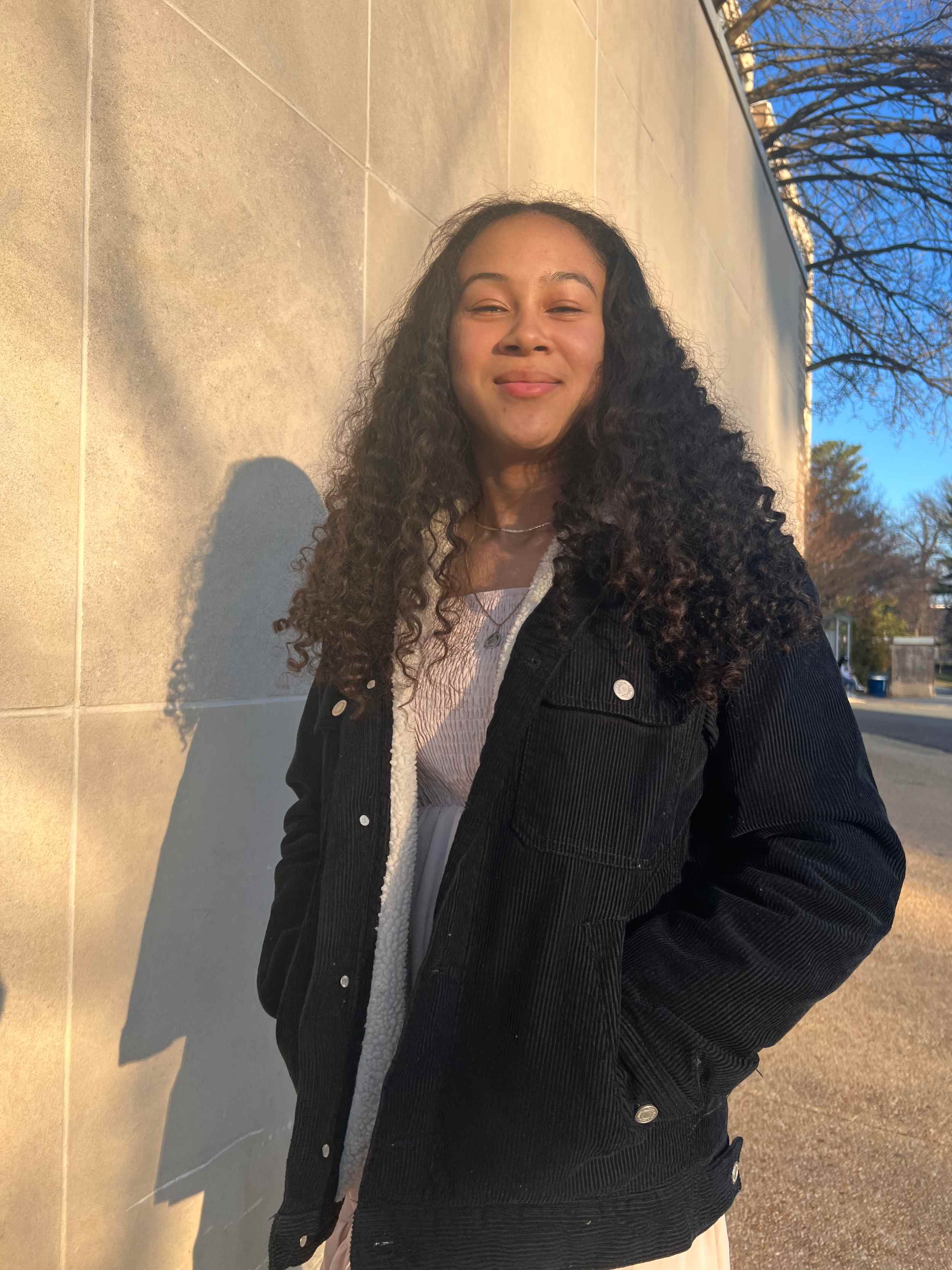 A photo of Savannah-Rae wearing a black jacket with her hands in her pockets standing in front of a white stone wall