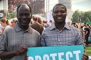 Didace Kamana stands on the National Mall with the Capitol Building in the background. He stands among scores of people, and he holds a sign saying Protect Our Common Home.