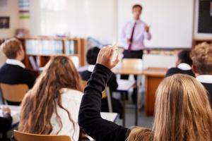 Young student seated in desk raises hand.